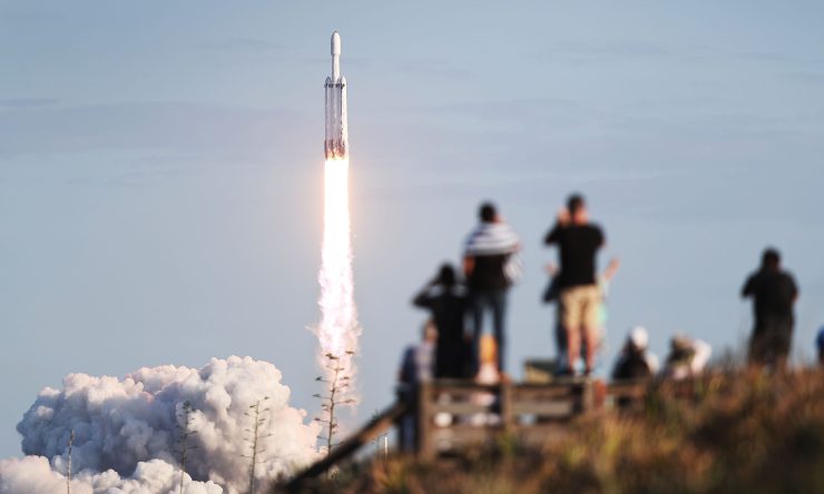 SpaceX launches a rocket in April 2019. In the foreground, several people stand and watch the rocket take off.