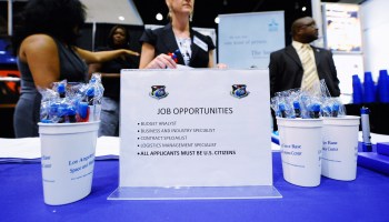 At a job fair, people stand behind a table with sign listing job opportunities and promotional pens in cups.