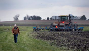 A farmer stands in a field near farm machinery that is depositing fertilizer into the soil.
