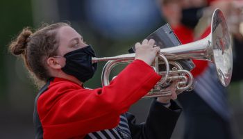 A high school marching band member plays a horn with a mask on.