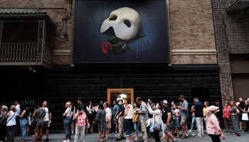 People in New York wait in line to see "Phantom of the Opera" under a banner with the mask logo.