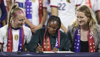 Three members of the U.S. women's soccer team sit at a table; Crystal Dunn, center, signs the equal pay contract.
