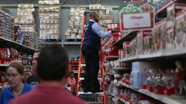 A Walmart worker on a ladder gets a holiday item off a top shelf for a customer.