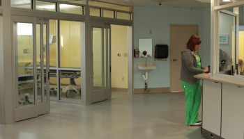 An emergency room hospital worker types at a nurses station, with patient rooms behind her.