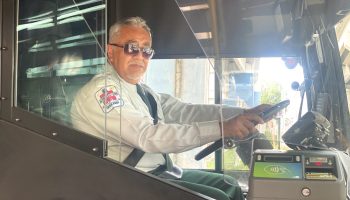 A bus operator sits in the drivers seat of a bus and turns to look at the camera.