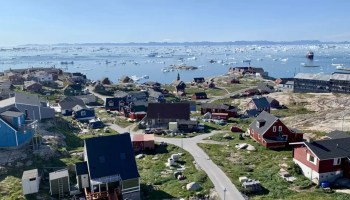 Steep-roofed homes painted red and gray dot the rolling green hills of Ilulissat, overlooking a bay filled with smaller icebergs.