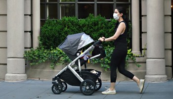 A woman wearing a protective mask and athletic wear pushes a stroller past a building with columns and greenery.