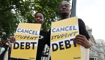 Two men hold yellow signs that say "Cancel student debt" in black letters at a student loan cancellation rally