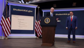 President Joe Biden gestures while standing behind a lectern with the presidential seal. Behind him on the right is Secretary of Education Miguel Cardonas; on the left, between American flags, is an enlarged view of StudentAid.gov.