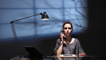 A woman sits at an office desk in a darkened room, lit by a desk lamp. She holds a phone to her ear, with a look of wariness on her face. On the wall behind her is the shadow of a tree's bare branches.
