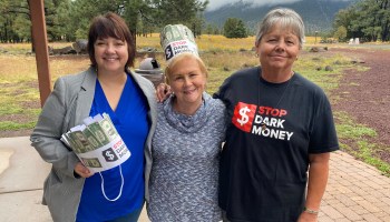 Three women in a park are holding or wearing items with the slogan Stop Dark Money.