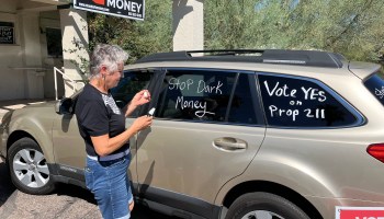 A woman stands by an SUV holding a white paint marker. She's written on the windows of the car "Stop Dark Money" and "Vote Yes on Prop 211."