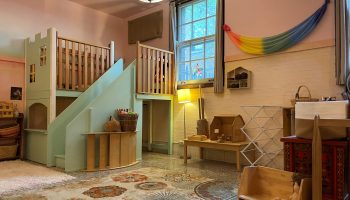 A brightly colored classroom at The Rose Garden Early Childhood Center has a castle-like playhouse with stairs leading to a top level. There's a rainbow-colored banner on a wall next to a window and a small wooden dollhouse on a table.