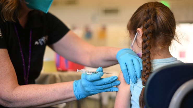 A nurse in gloves administers a pediatric dose of the COVID-19 vaccine to a girl at a Los Angeles clinic. The girl is sitting in a chair, her back to the camera. Her long, brown braid hangs beside the earpiece of her glasses.