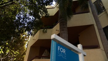 A "for sale" sign outside a townhouse-style building shaded by a tree in Los Angeles.