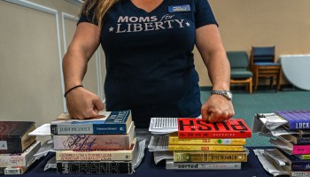 A woman wearing a T-shirt that reads Moms for Liberty stands in front of stacks of books.