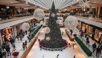 A shopping mall at Christmastime, with a huge Christmas tree in the middle of the floor. and glittery decorations hanging from the ceiling.