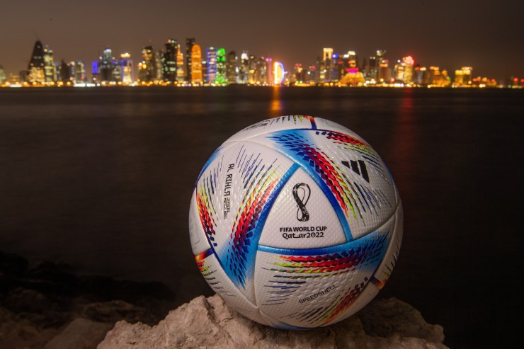 A soccer ball with the text "FIFA World Cup, Qatar 2022" sits on a rock in front of a panoramic view of the Doha skyline.