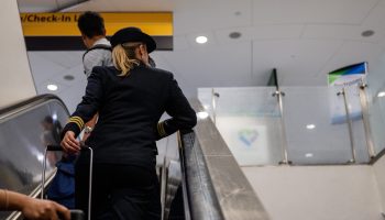 A pilot and travelers ride an escalator up at an airport.