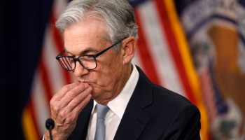 Fed Chair Jerome Powell bows his head slightly, resting his chin on his fingers, while answering questions at a press conference. Behind him is the American flag.