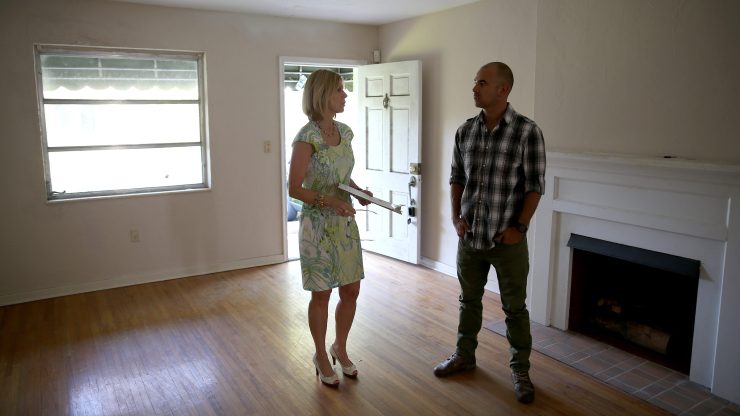 A man stands in an empty house with a real estate agent. The front door is open, and the room has a fireplace and window.