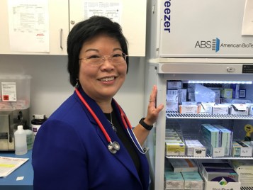 Sandy Chung stands next to an open cooler showing shelves filled with boxes of various vaccines.