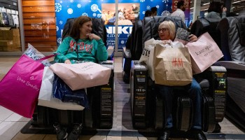 Two women looking at each other sit in massage chairs in a mall, overloaded with shopping bags.