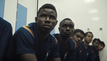 A row of African and Asian migrant worker soccer players sit on a bench in a changing room before a big match.