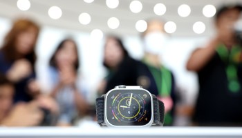 A new Apple Watch is displayed on a table with people looking at the device in the background.