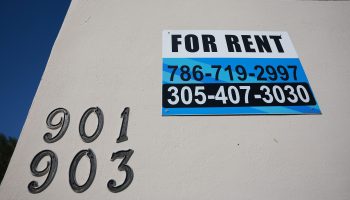 A blue "For Rent" sign on the outside of a cream-colored building.