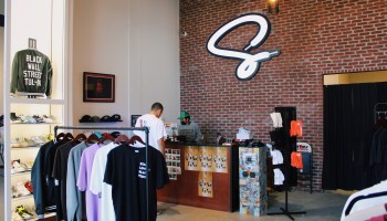 A giant cursive "S" formed from a shoe lace hangs on a brick wall behind the counter at Silhouette Sneakers. A person in a green hat helps a customer at the register. In the foreground, T-shirts hang from a rack and shoes line a wall.