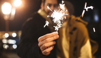 A couple wearing coats at night lean in close to one another, while the bearded man holds out a sparkler with one hand.