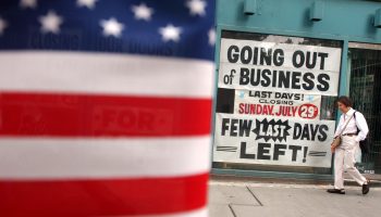 A woman walks past a "Going out of business" sign with an American flag partially seen in the foreground.