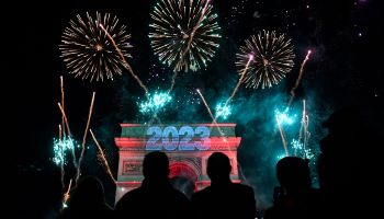 Fireworks explode above a colorfully lit Arc de Triomphe with "2023" projected on the building. Silhouettes of people watching are in the foreground.