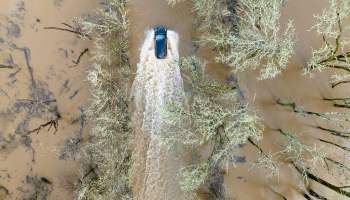 An aerial view of a blue vehicle driving through a muddy, flooded street surrounded by trees.