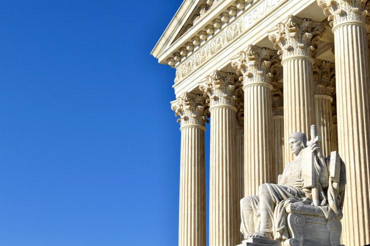 A seated male figure carved of marble sits in front of the columns in front of the Supreme Court. The white marble is contrasted against a blue sky.