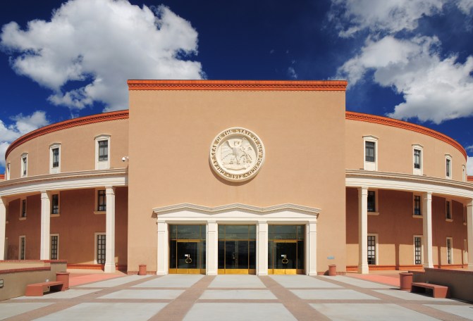 The New Mexico State Capitol Building is seen against clouds in a blue sky. 