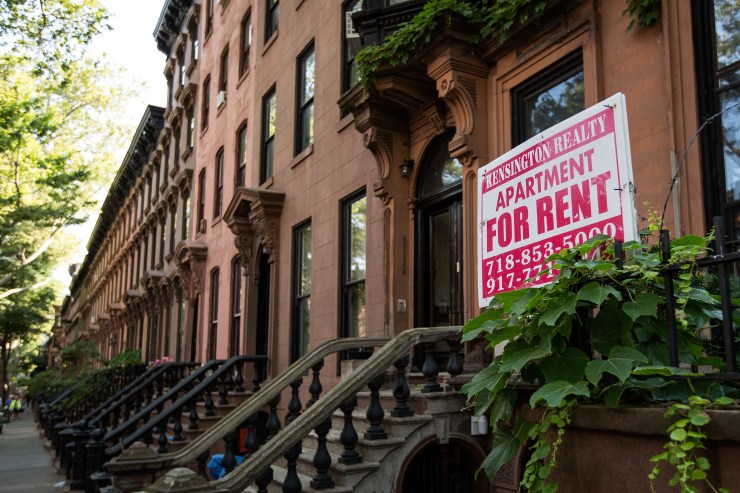 A row of red-stone apartments in Brooklyn with a "For Rent" sign in front.