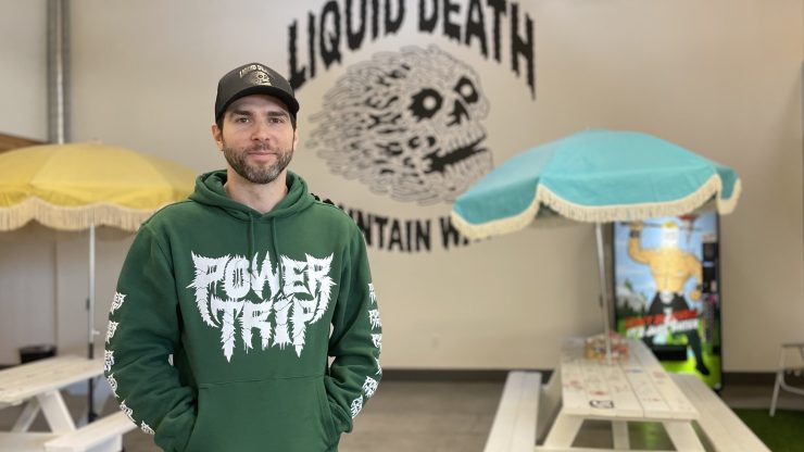 Mike Cessario, a bearded man, stands in a room with picnic tables and sun umbrellas, with Liquid Death's skull logo on the back wall. Cessario is wearing a green hoodie that says "Power Trip" and a Liquid Death baseball hat.
