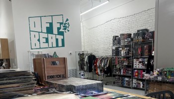 Vinyl albums are stacked and sit in bins on a table at OffBeat. Behind it, the store logo is outlined in green on a white wall, next to T-shirts and other items for sale.