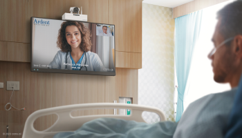 A patient sits in a hospital bed while a nurse is streamed on an adjacent television.