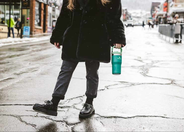 A woman stands in the middle of a street with a black coat, pants and boots. She is holding a teal-colored Stanley tumbler.