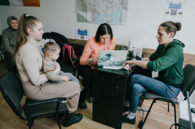 Mo Hornik sits at a table pointing at paper work with a Ukrainian refugee family — a mother, a young adult daughter and an infant.