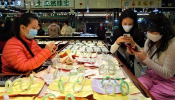 A jade seller on the left, with black hair, a blue face mask and orange jacket, sits at a table staring at her cell phone. Across the table, two other women with black hair and face masks also sit on their phones. On the table are perfectly round jade bracelets.