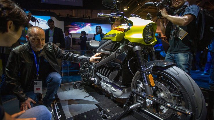 In a convention hall, people gather around a black and gold Harley-Davidson electric motorcycle on a stand.