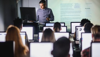 A middle-aged white man with salt-and-pepper hair stands at the front of a classroom behind a computer. On a screen projector behind him, a block of text is shown. In the foreground, rows of students face the instructor, each with a computer screen lit up in front of them.