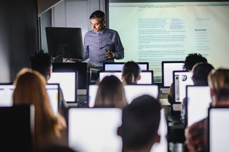 A middle-aged white man with salt-and-pepper hair stands at the front of a classroom behind a computer. On a screen projector behind him, a block of text is shown. In the foreground, rows of students face the instructor, each with a computer screen lit up in front of them.