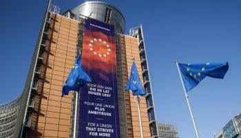 Three European Union flags — blue field with a ring of 12 yellow stars — fly in front of the European Commission in Brussels, Belgium.