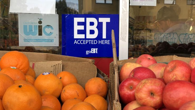 A sign noting the acceptance of electronic benefit transfer (EBT) cards is displayed in a shop window behind two cardboard boxes, one filled with oranges, the other filled with oranges.