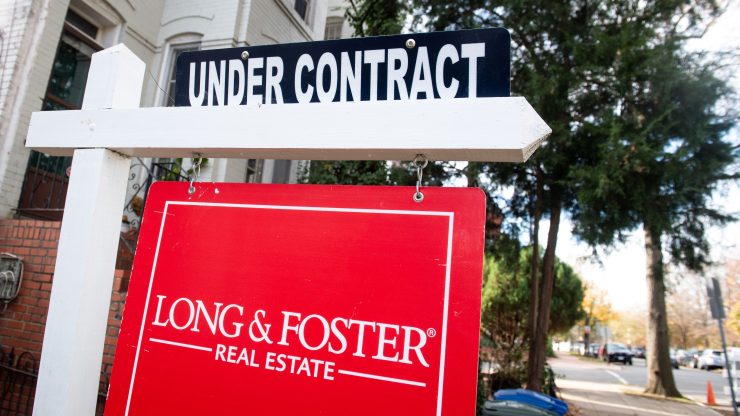 A red Long & Foster real estate sign hangs from a white post. On top is a smaller sign reading: Under Contract. In the background is a sidewalk and street lined with mature, leafy trees.